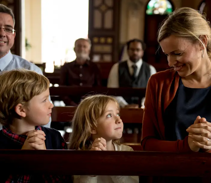 Familia integrada por papá, mamá y dos hijos en una iglesia católica en posición de oración, arrodillados con las manos unidas.